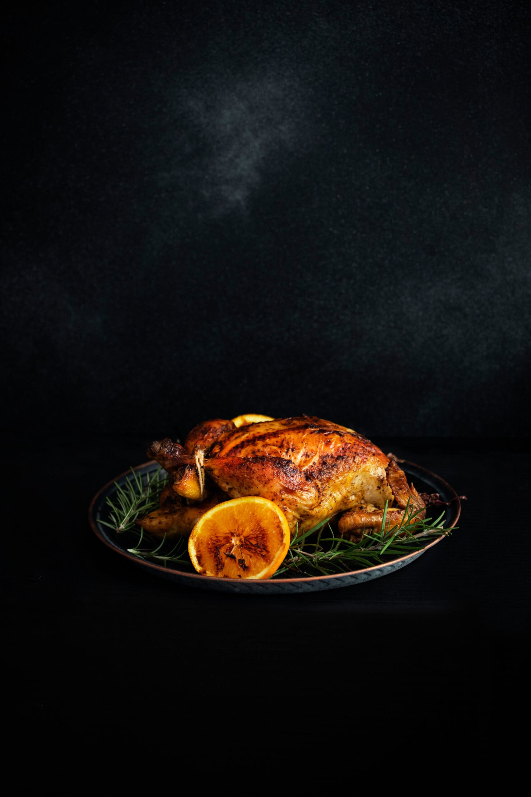 Baked whole chicken with oranges and rosemary. Tray with a festive Christmas dish on a black table and black background. Low key. Vertical. Copyspace